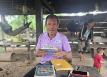 ប្រធានសហគមន៍នេសាទ លោក ខាន់ ប៊ុនទី ជាជនជាតិទំពួននៅភូមិកែងសាន់ ឃុំសេដា ស្រុកលំផាត់ ខេត្តរតនគិរី កំពុងអង្គុយកាន់សៀវភៅនៅក្រោមផ្ទះកាលពីថ្ងៃទី២៦ ខែកក្កដា ឆ្នាំ២០២៥។ (សាមន្ត ដារ៉ា)